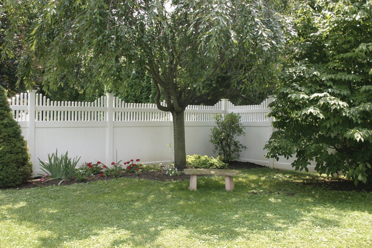 View of the corner of a white semi privacy fence surround a yard with green grass, tall trees, and a flower bed in the corner