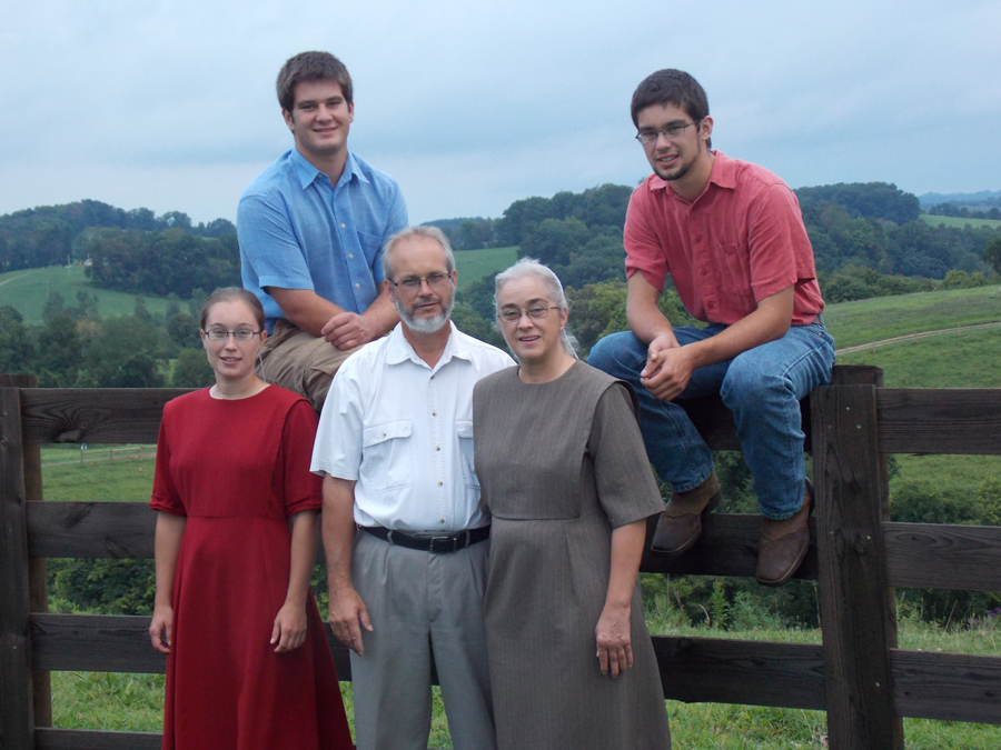 A family of five posing for a picture with three standing together and two sitting behind on a wooden fence, with a wide open field as a backdrop