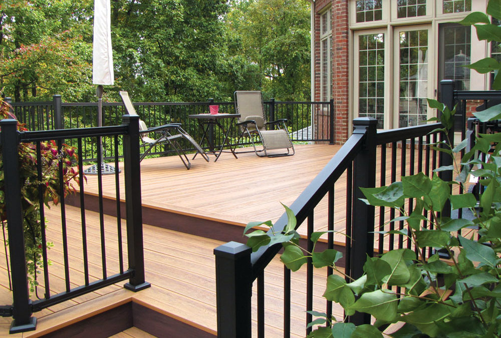 A spacious wooden deck with black railing attached to the back of a house surrounded by greenery