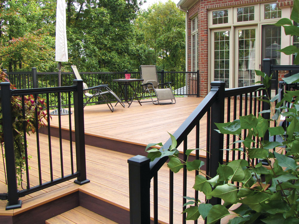 A spacious wooden deck with black railing attached to the back of a house surrounded by greenery