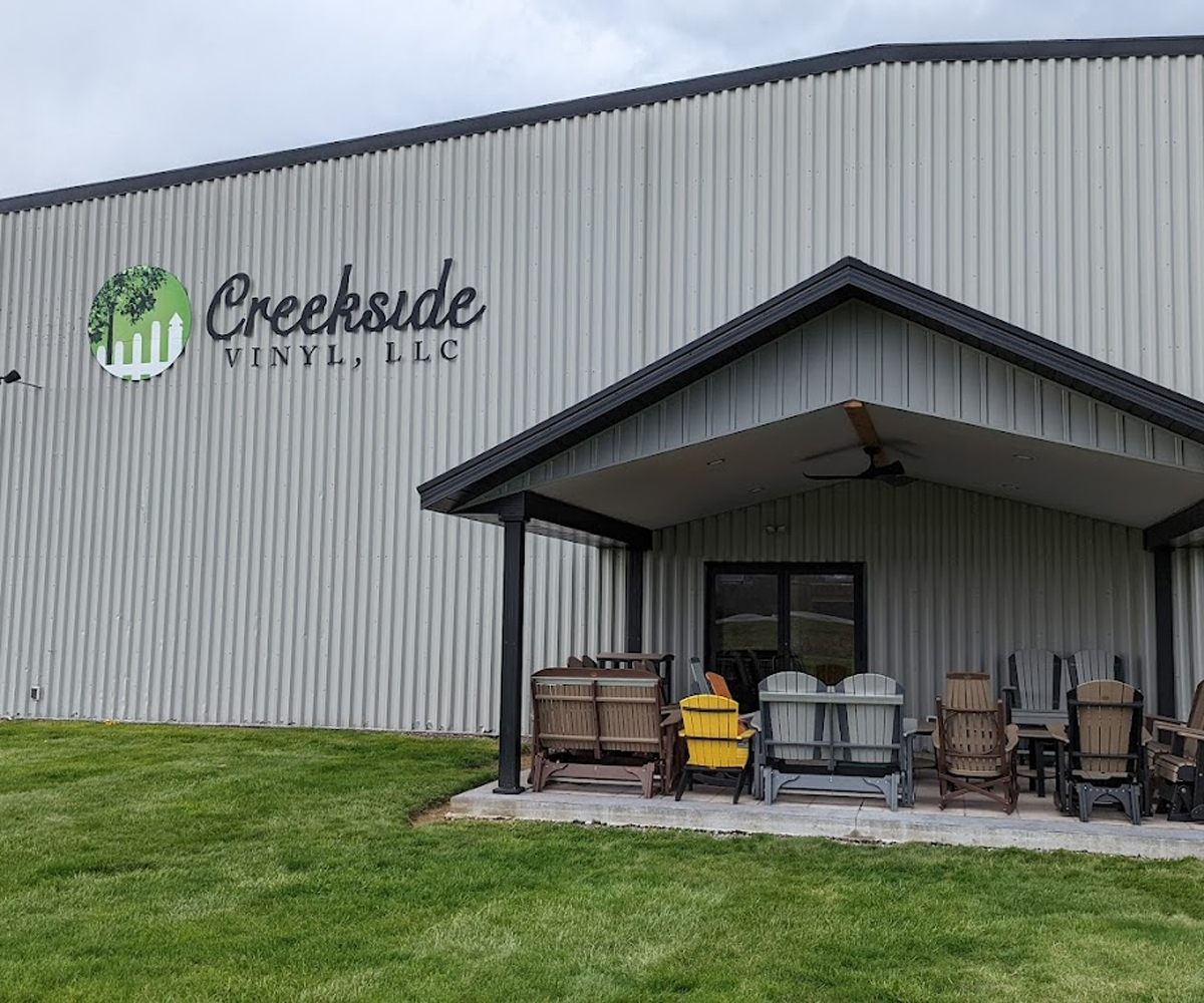 Exterior of the Creekside Vinyl, LLC metal building with a large company logo and an array of poly chairs under the covered porch. Contact Us and find aluminum or vinyl railing and poly furniture for your backyard.